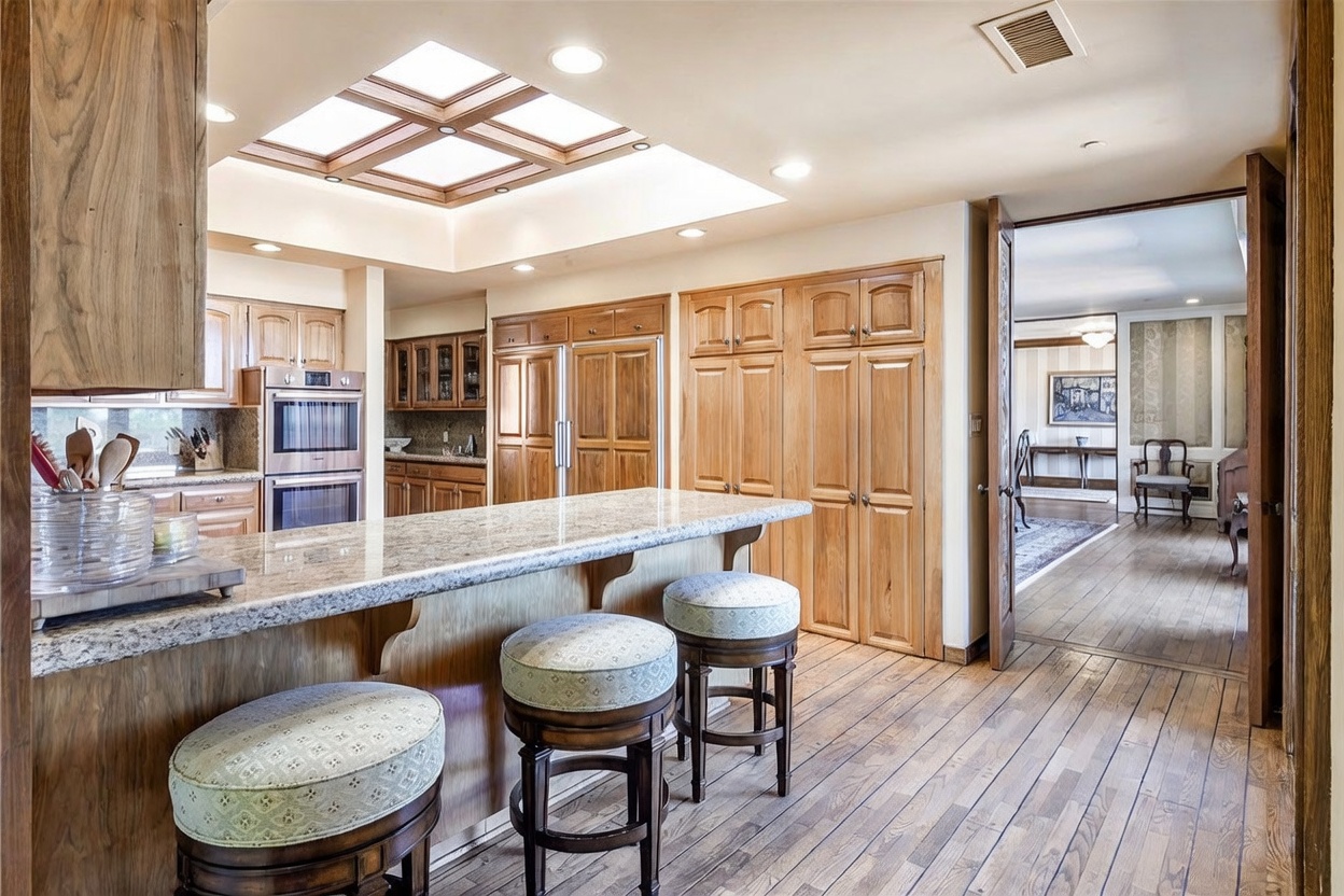 This kitchen features a large island with a stone countertop and three stools, set against a backdrop of light wood cabinetry and built-in appliances. A prominent skylight with a wood-trimmed frame provides natural light from above, while a doorway leads into an adjacent room with wood flooring. The space has a traditional aesthetic with warm wood tones and a functional layout.
