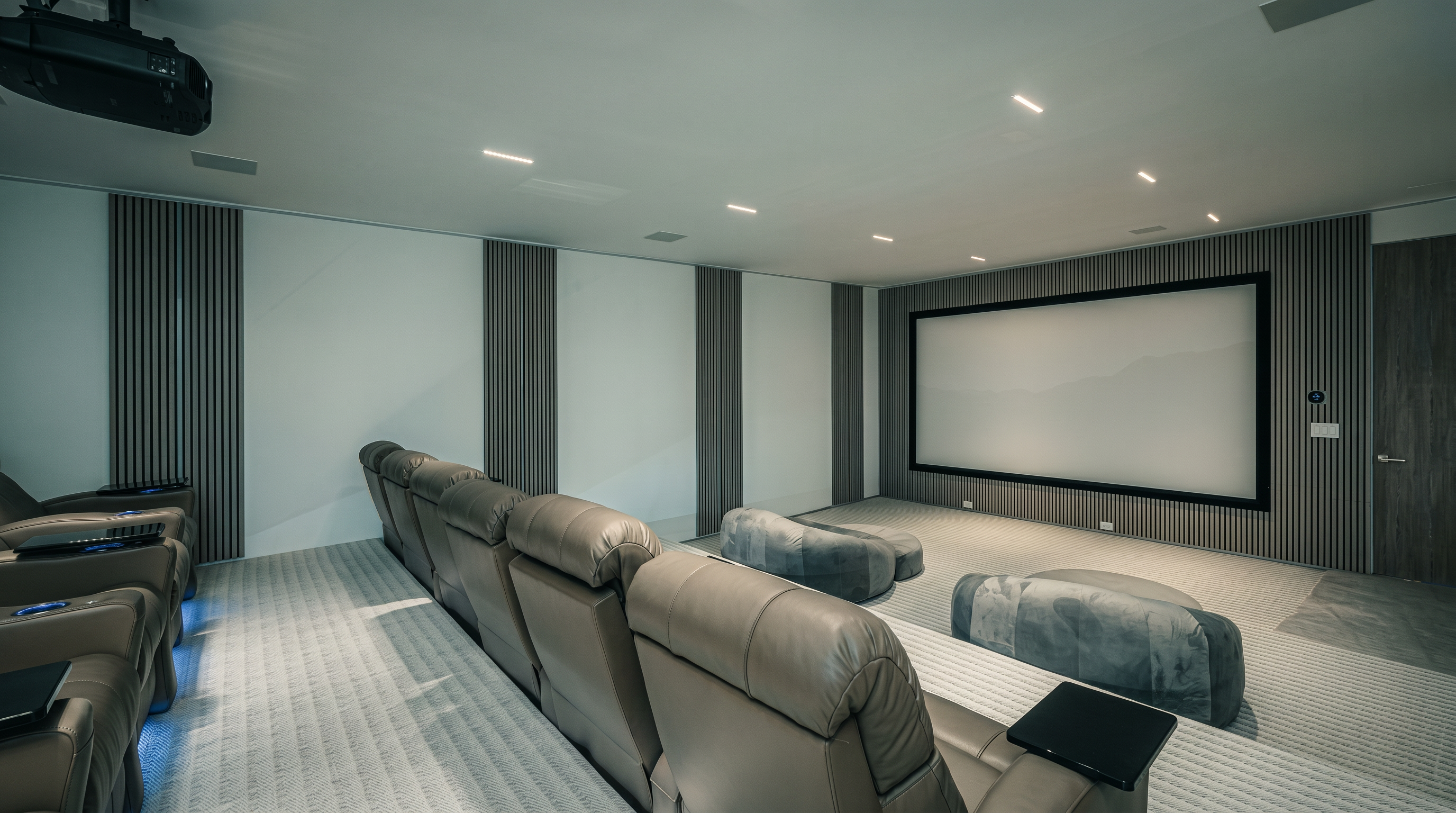This dedicated home theater room features tiered rows of plush, light-colored leather reclining seats facing a large projection screen. The walls are accented with vertical wood-slat paneling, and the room is equipped with a ceiling-mounted projector and recessed lighting. The space is designed for an immersive cinematic experience, complete with additional floor-level seating cushions near the screen.