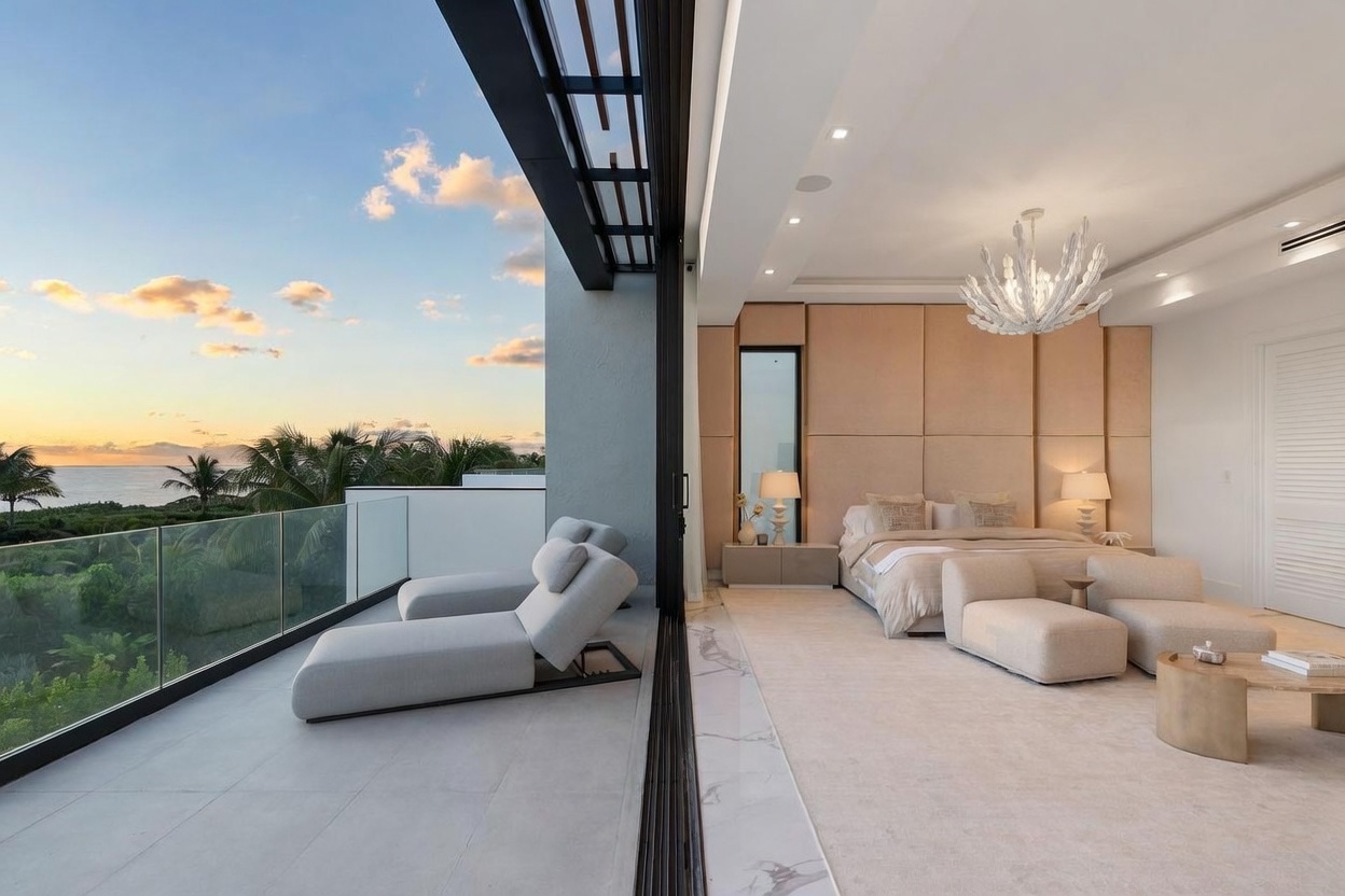 This luxurious primary bedroom features a seamless transition to a private balcony through expansive floor-to-ceiling sliding glass doors. The room is anchored by a large bed with a neutral-toned, paneled headboard wall and is illuminated by a striking, sculptural chandelier. The balcony offers a serene outdoor lounge area with two chaise lounges, providing a panoramic view of the ocean and tropical landscape at sunset.