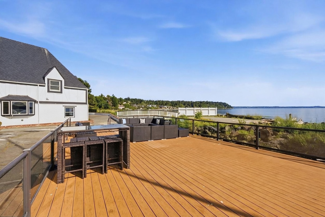This expansive, elevated wooden deck offers a panoramic view of the water and distant shoreline. The space is furnished with a modern outdoor sectional sofa and a high-top dining table, all enclosed by a sleek glass railing that maintains an unobstructed view. The scene is captured from a slightly elevated perspective, highlighting the seamless transition between the home's exterior and the natural coastal landscape.