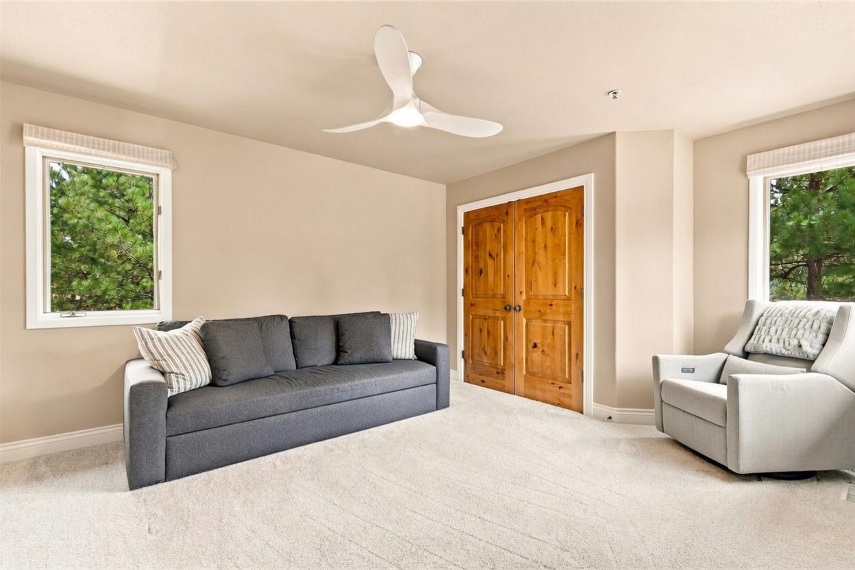 This bright and neutral bedroom features a dark gray sofa and a light gray recliner, creating a comfortable seating area. The room is accented by two windows that provide natural light and views of the surrounding trees, while a modern white ceiling fan hangs centrally. A set of warm-toned wooden double doors adds a touch of traditional character to the otherwise minimalist space.
