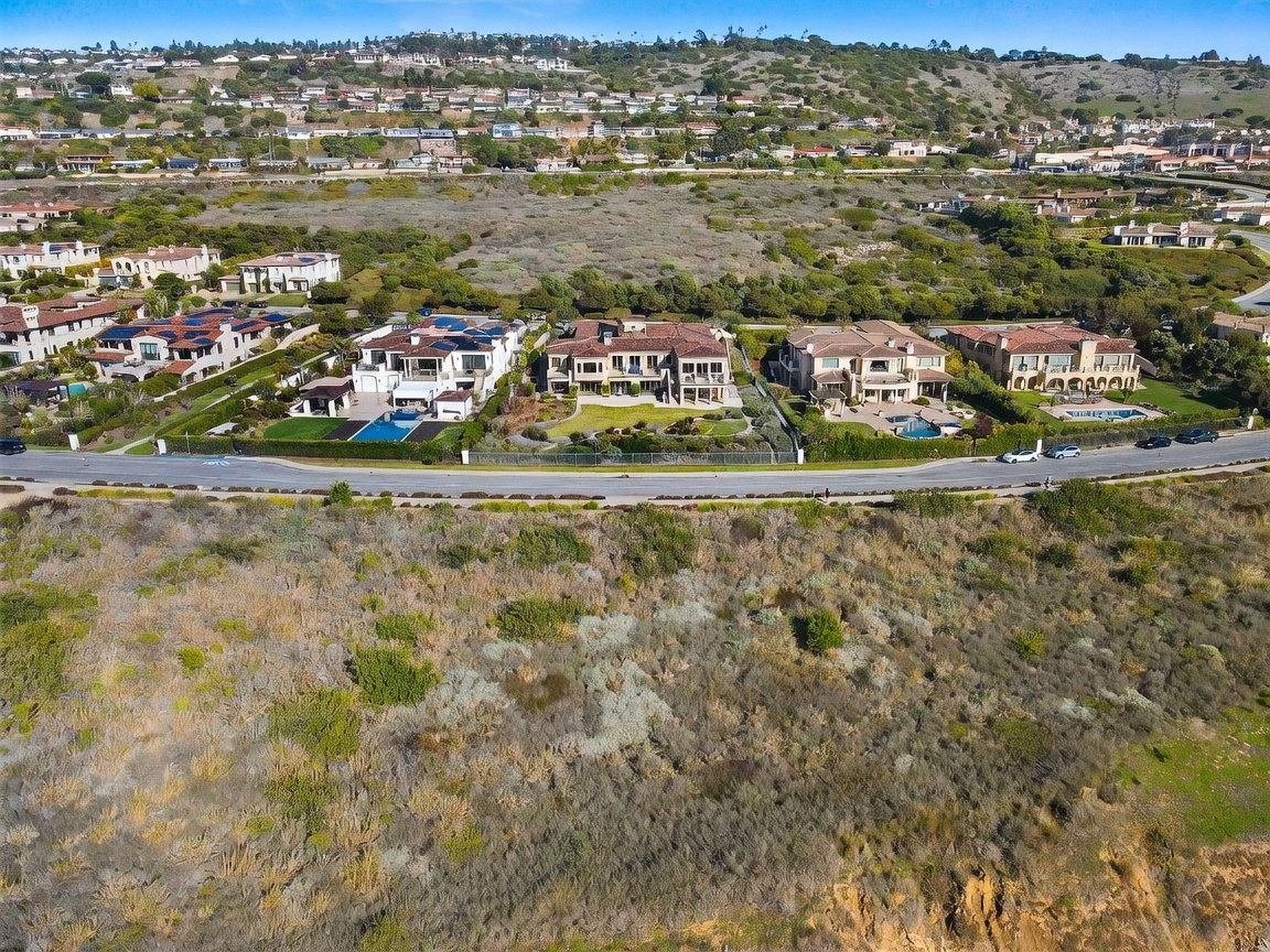 This high-angle aerial view captures a row of luxury Mediterranean-style homes situated along a coastal bluff. The foreground features a winding pedestrian trail cutting through natural, brush-covered terrain, while the homes showcase terracotta-tiled roofs, private swimming pools, and manicured lawns. The perspective emphasizes the expansive, scenic location and the proximity of the residences to the open landscape.