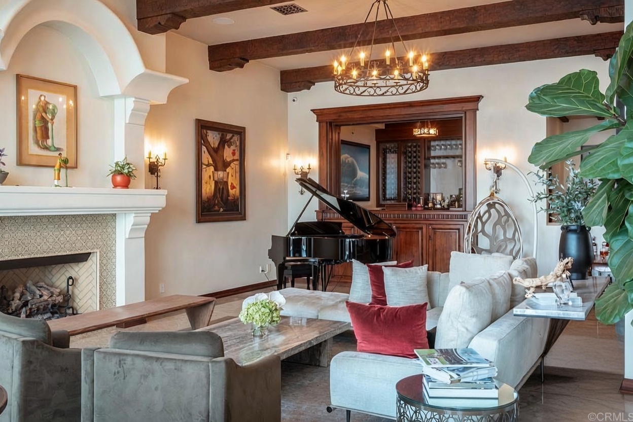 This elegant living room features a grand piano as a central focal point, set against a backdrop of dark wood cabinetry and a built-in bar area. The space is defined by exposed wooden ceiling beams, a fireplace with a decorative mantel, and a comfortable seating arrangement with plush sofas and armchairs. The warm lighting from the chandelier and wall sconces creates an inviting and sophisticated atmosphere, perfect for entertaining.