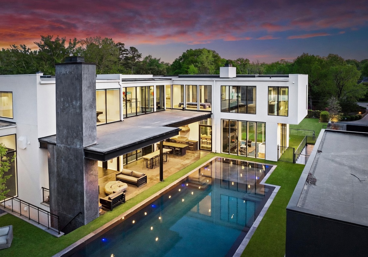 This aerial perspective captures a modern, multi-level residence characterized by clean white lines, expansive glass walls, and a prominent dark stone chimney breast. The home features a covered outdoor living area with a dining space and lounge, which transitions seamlessly to a rectangular swimming pool set within a manicured lawn. The composition highlights the architectural symmetry and the integration of indoor-outdoor living spaces during a vibrant sunset.