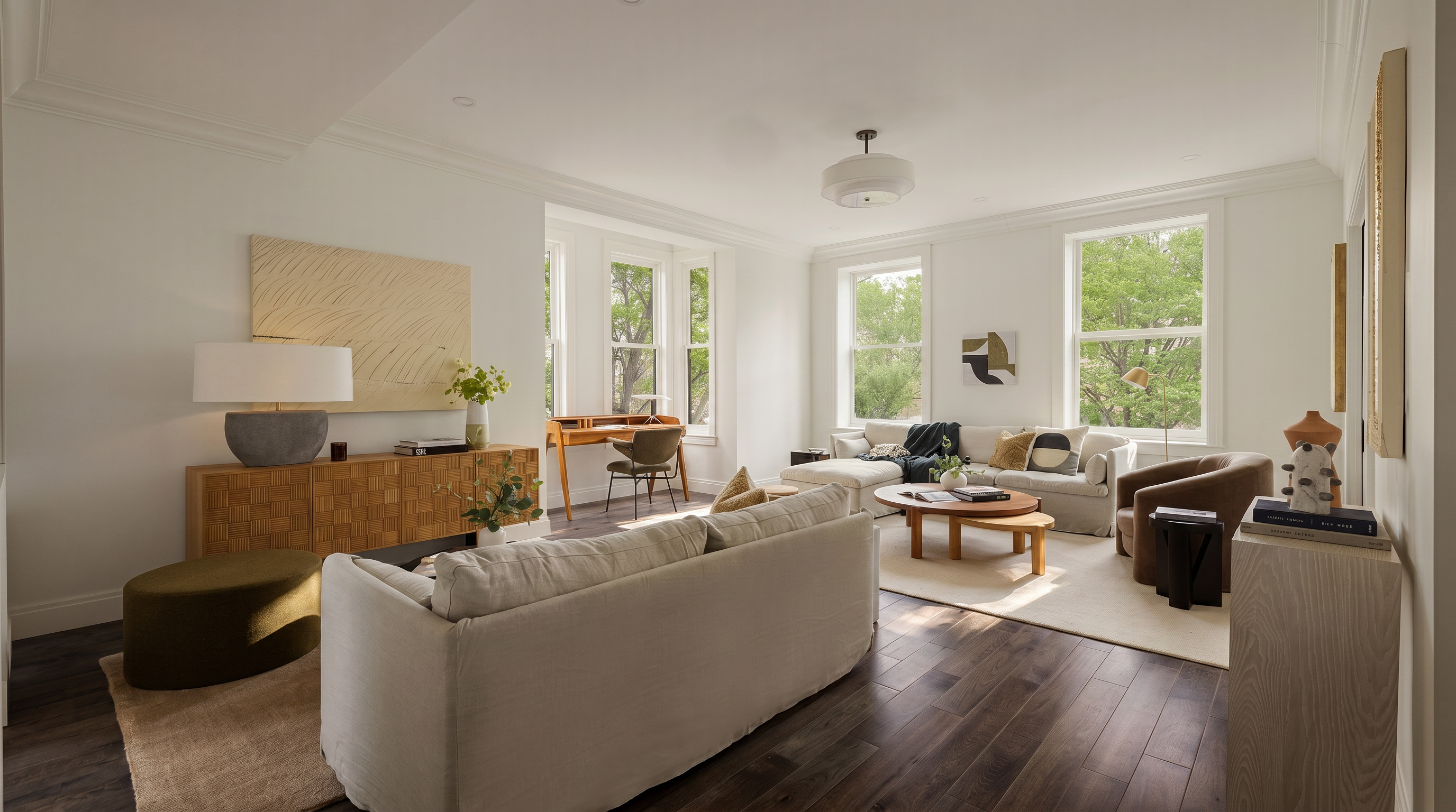 This bright and airy living room features a sophisticated, neutral color palette with warm wood flooring and high ceilings. The space is furnished with a plush cream sofa, a textured sideboard, and a cozy seating area near large windows that invite ample natural light. The open-concept layout seamlessly integrates a dedicated workspace, creating a versatile and inviting atmosphere perfect for modern living.