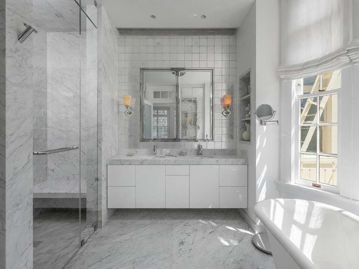 This elegant primary bathroom features extensive use of light-toned stone on the floors and shower walls, creating a bright and cohesive aesthetic. The space includes a double vanity with a sleek white cabinet, a glass-enclosed walk-in shower, and a freestanding soaking tub positioned near a window. A mirrored medicine cabinet and wall-mounted sconces add a touch of sophistication to the clean, modern design.