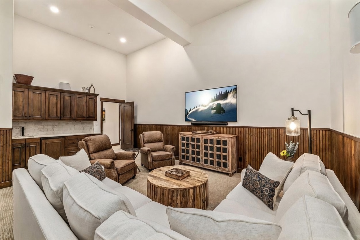 This spacious living room features high ceilings and a warm, rustic aesthetic defined by wood-paneled wainscoting. The room is furnished with a large, light-colored sectional sofa, two leather recliners, and a round wooden coffee table, all centered around a wall-mounted television. A built-in wet bar with dark cabinetry and a countertop provides a convenient space for entertaining, while a floor lamp adds soft lighting to the cozy seating area.