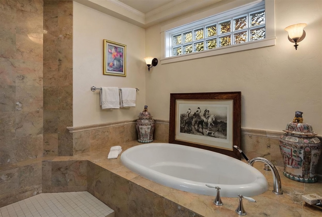 This elegant bathroom features a deep soaking tub set within a stone-tiled deck, complemented by a matching stone-tiled shower surround. The space is adorned with decorative porcelain jars, framed artwork, and a transom window composed of glass blocks that allows for soft, diffused natural light. The warm color palette and classic fixtures create a sophisticated and tranquil atmosphere.