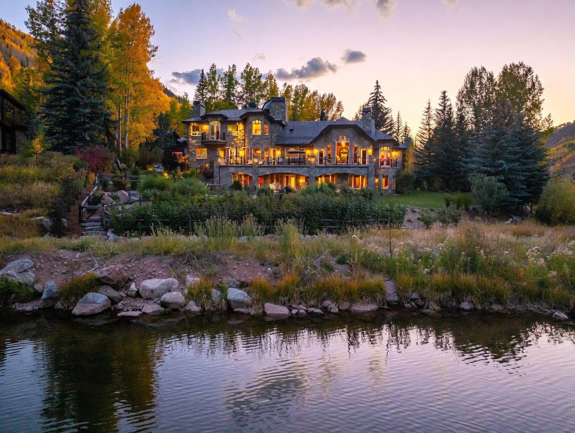 This stunning multi-story stone estate is captured from a low-angle perspective across a tranquil pond, highlighting its grand architectural presence at dusk. The home features a sprawling rear terrace, multiple balconies, and warm interior lighting that glows through numerous large windows, creating an inviting and luxurious atmosphere. Surrounded by lush trees with autumn foliage and a manicured lawn, the property exudes a serene, high-end mountain retreat aesthetic.