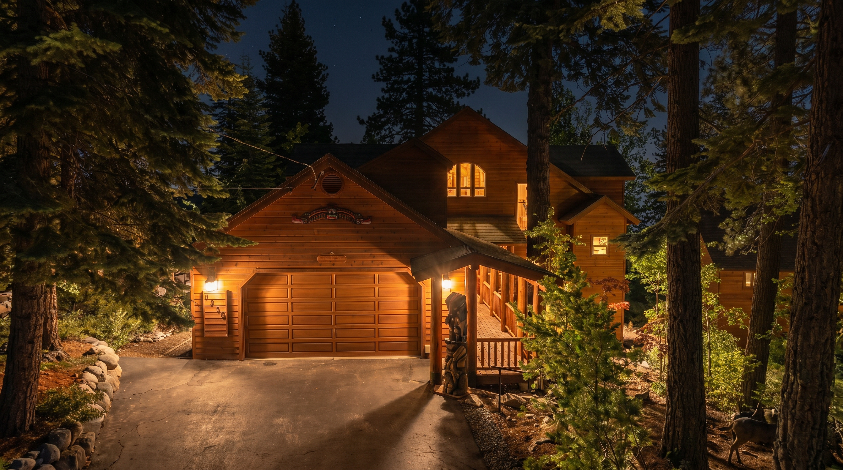 This charming, multi-level mountain cabin features warm-toned wood siding, a prominent two-car garage, and a welcoming covered porch adorned with a decorative totem pole. Set in a wooded landscape at night, the home is illuminated by warm exterior lighting that highlights its rustic architectural style and cozy, forested appeal. It provides a classic, inviting appearance typical of a high-end mountain retreat.