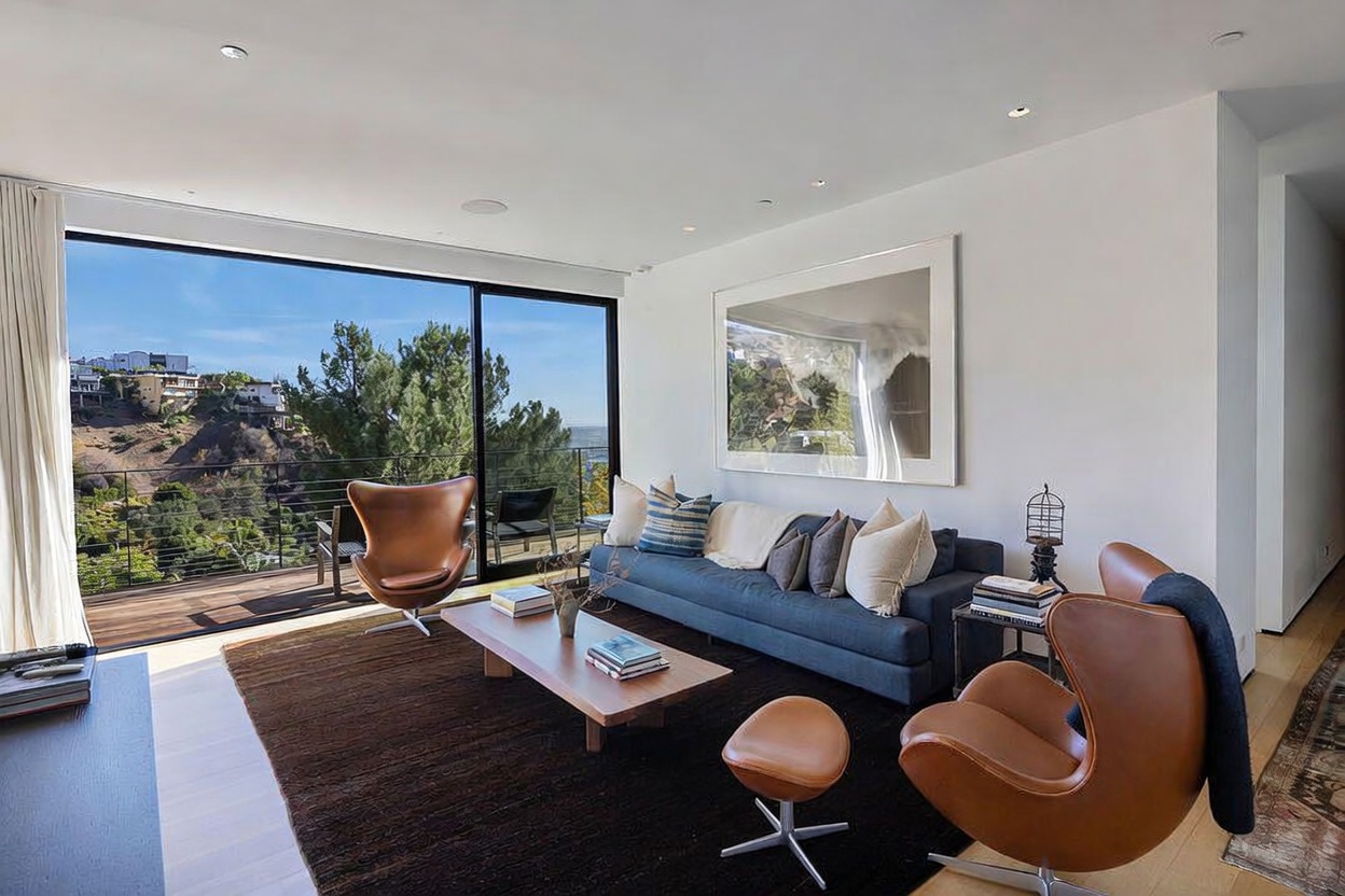 This modern living room features a sophisticated design with a dark blue sofa, two iconic leather lounge chairs, and a matching ottoman arranged on a large dark area rug. The space is anchored by a low-profile wooden coffee table and illuminated by recessed lighting, while a large piece of wall art adds a focal point to the room. Expansive floor-to-ceiling glass doors provide a seamless transition to an outdoor balcony, offering panoramic views of the surrounding hillside landscape.