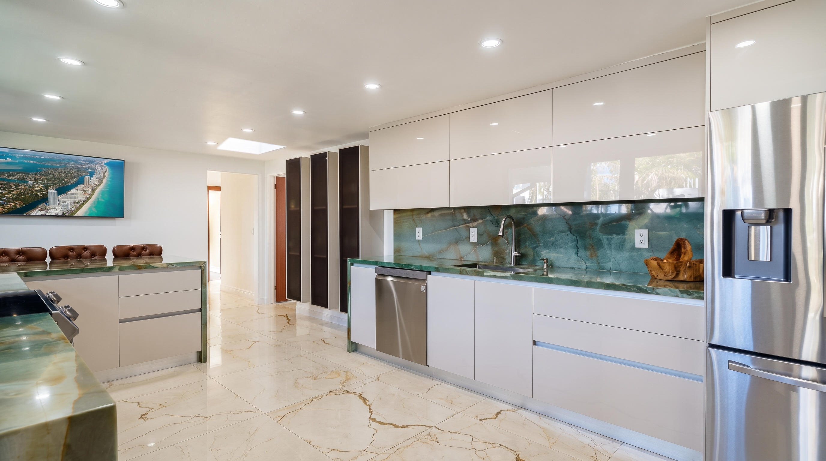 This modern kitchen features sleek, high-gloss white cabinetry paired with a striking green stone backsplash and matching countertops. The space is illuminated by recessed ceiling lights and includes stainless steel appliances, such as a dishwasher and a large refrigerator. The floor is finished with light-colored, veined stone tiles, and a television is mounted on the wall, providing a view into an adjacent living area.