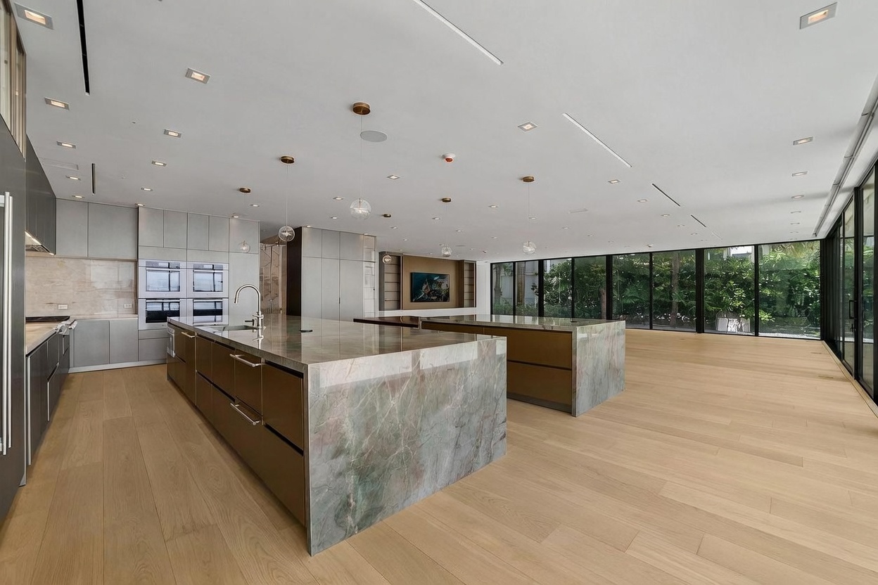 This expansive, modern kitchen features two large islands with stone countertops and dark cabinetry, set against a backdrop of light wood flooring. The space is illuminated by minimalist pendant lights and recessed lighting, while floor-to-ceiling glass walls provide an unobstructed view of the lush greenery outside. The open-concept design creates a seamless flow, emphasizing a sleek, high-end aesthetic suitable for contemporary living.