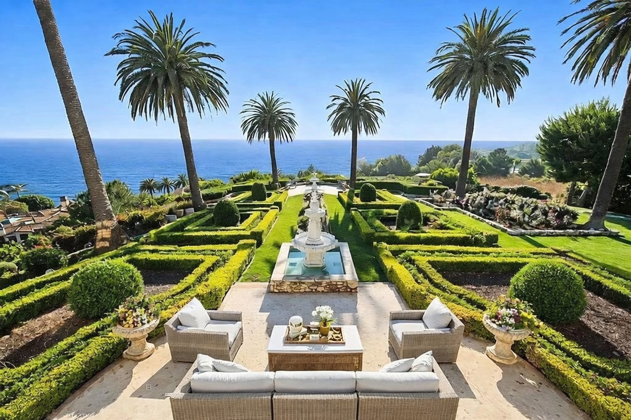 This elevated perspective captures a meticulously manicured formal garden featuring geometric boxwood hedges and a central tiered stone fountain. The outdoor living space is furnished with a comfortable sofa and armchairs, creating a serene lounge area that overlooks the expansive blue ocean. Tall palm trees frame the horizon, enhancing the luxurious, resort-like atmosphere of this coastal property.