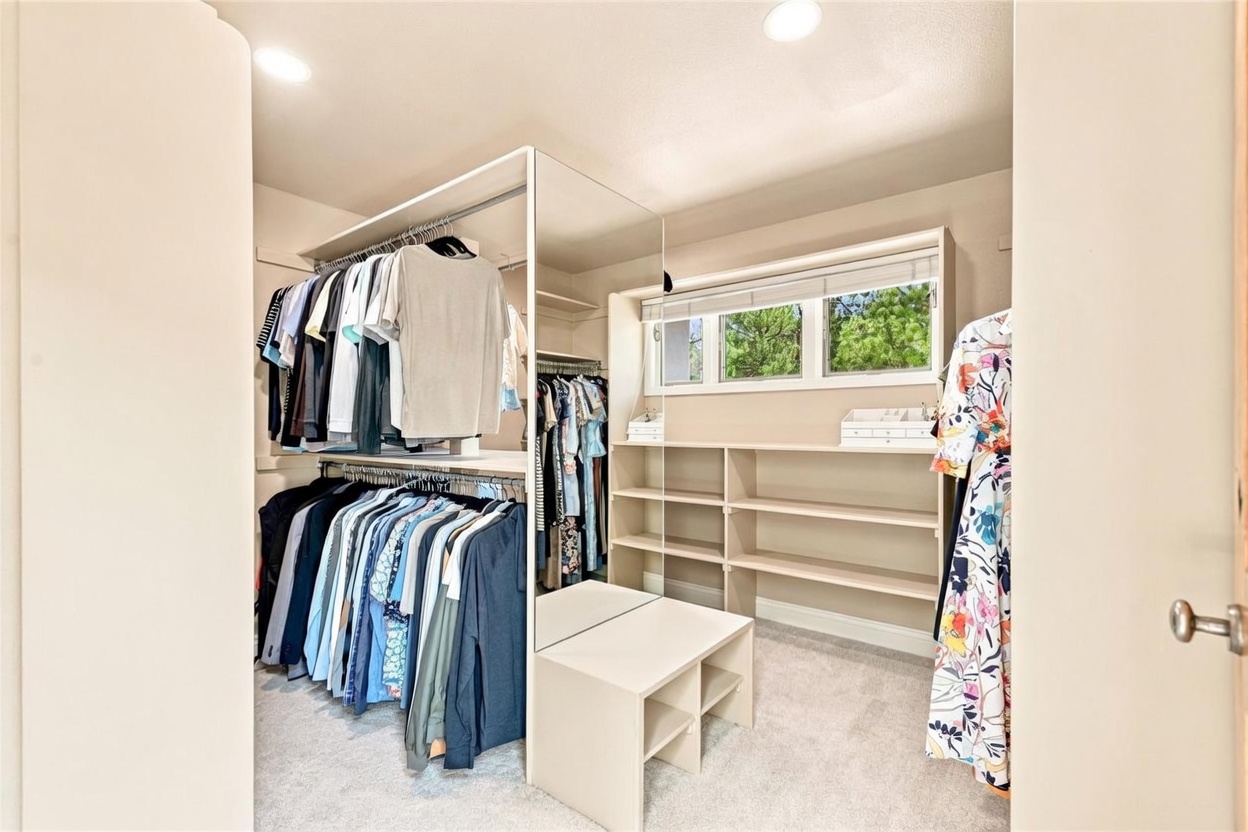 This organized walk-in closet features a dual-level hanging rod system on the left and a central mirrored partition that separates the space. To the right, a window with blinds provides natural light above a series of open shelving units, while a small bench with storage cubbies sits in the center. The room is finished with neutral-toned walls and light-colored carpeting, creating a functional and tidy storage area.