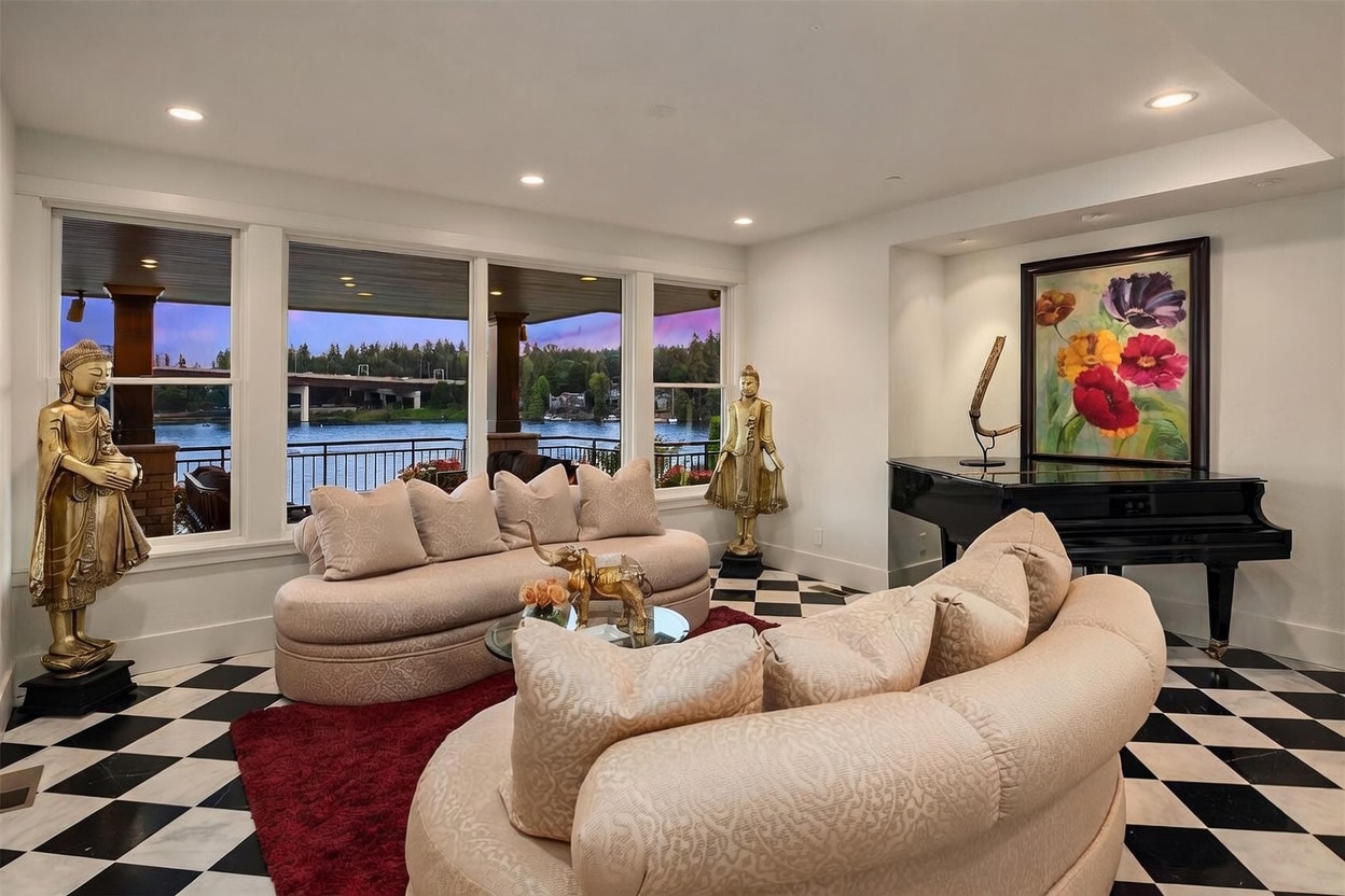 This elegant living room features a striking black-and-white checkered floor and two curved, light-toned sofas arranged around a glass coffee table. Large windows provide an expansive view of the water and a bridge, while decorative gold-toned statues and a vibrant floral painting add a sophisticated, artistic touch. A grand piano sits in the corner, completing the luxurious and refined atmosphere of the space.