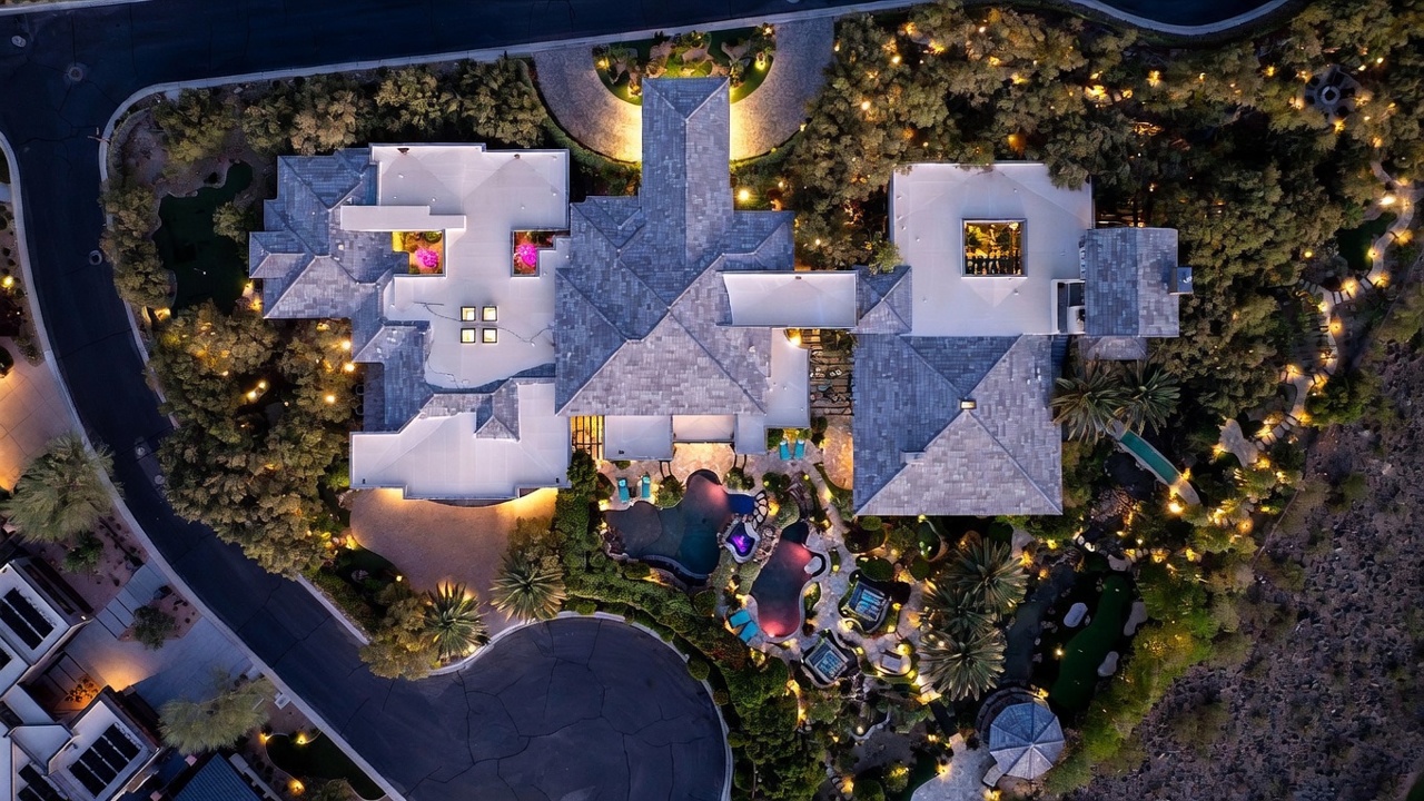 This high-angle aerial view captures a sprawling luxury estate at twilight, showcasing its complex roofline and extensive outdoor living spaces. The property features a multi-section swimming pool, lush landscaping with mature palm trees, and a private putting green, all illuminated by warm landscape lighting. The perspective highlights the expansive layout of the residence and its integration into the surrounding desert-inspired terrain.
