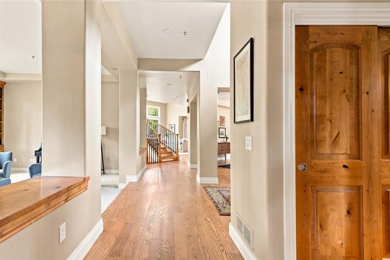 This interior view captures a bright, open hallway featuring light wood flooring and neutral-toned walls. A wooden staircase with black metal balusters serves as a focal point in the background, while a large wooden door on the right adds a warm, traditional touch. The perspective looks down the main corridor, highlighting the home's spacious layout and transition between living areas.