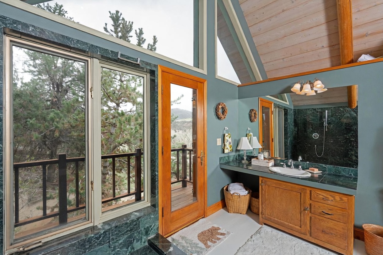 This bathroom features a vanity with a stone countertop and a wood cabinet, complemented by a large mirror and a wall-mounted light fixture. A glass door provides access to an outdoor deck, while large windows offer expansive views of the surrounding trees and landscape. The room is finished with stone accents and a vaulted ceiling, creating a rustic yet refined atmosphere.