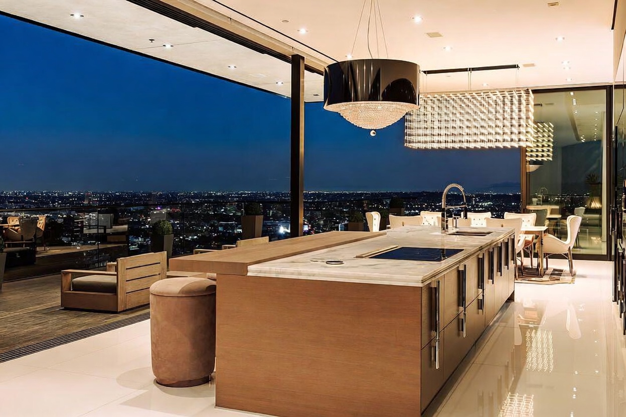 This ultra-modern, high-end kitchen features a massive marble-topped island with integrated appliances and a sleek wooden base, seamlessly blending into an expansive outdoor terrace. The space is illuminated by a dramatic, large-scale crystal chandelier and recessed lighting, while floor-to-ceiling glass walls offer a breathtaking, panoramic view of the city lights at night. The open-concept design creates a sophisticated, luxurious atmosphere perfect for high-end entertaining.
