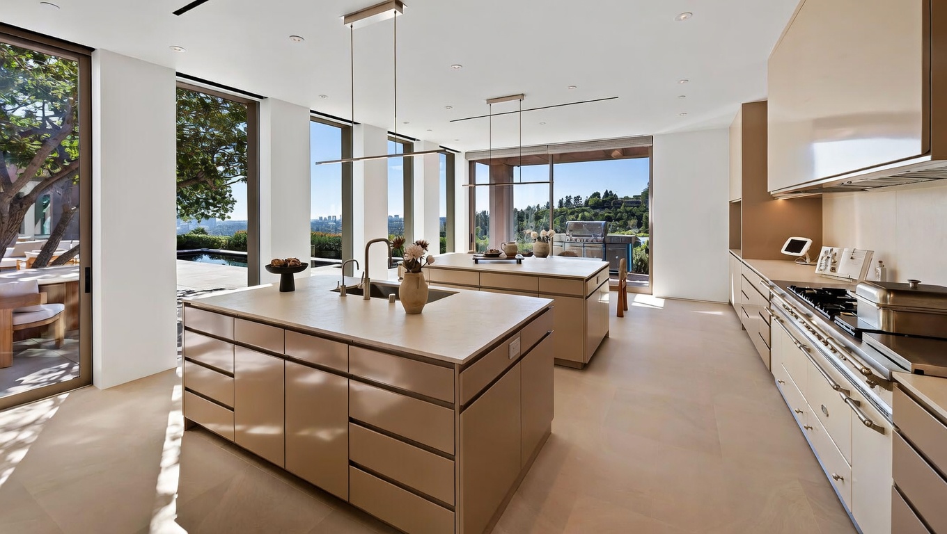 This expansive, modern kitchen features a minimalist design with sleek, light-tan cabinetry and dual large islands topped with stone countertops. The space is flooded with natural light from floor-to-ceiling glass doors that open to an outdoor patio and pool area, creating a seamless indoor-outdoor flow. The aesthetic is sophisticated and airy, emphasizing clean lines and high-end finishes suitable for luxury living.