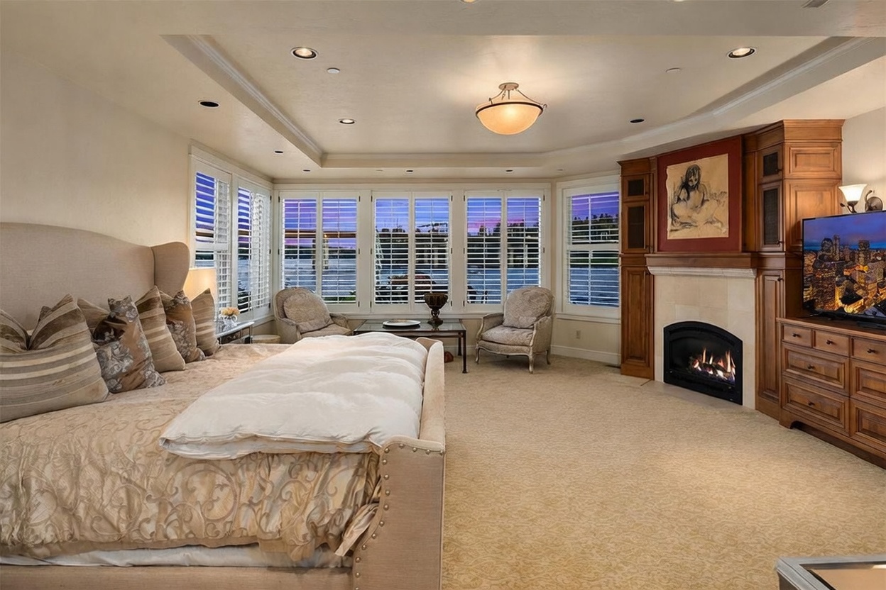 This spacious primary bedroom features a large, plush bed with patterned bedding and a neutral-toned headboard. The room is anchored by a fireplace with a wood-trimmed surround and a television mounted above, while a seating area with two armchairs is positioned in front of large windows overlooking the water. The space is finished with wall-to-wall carpeting and recessed lighting, creating a serene and luxurious atmosphere.
