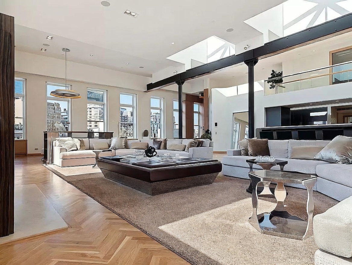 This expansive, open-concept living area features high ceilings with exposed structural beams and large windows that flood the space with natural light. The room is anchored by a massive, dark-toned central coffee table surrounded by plush, light-colored sectional seating on a textured area rug. The herringbone-patterned wood flooring adds a classic touch to the modern, industrial-inspired architecture.