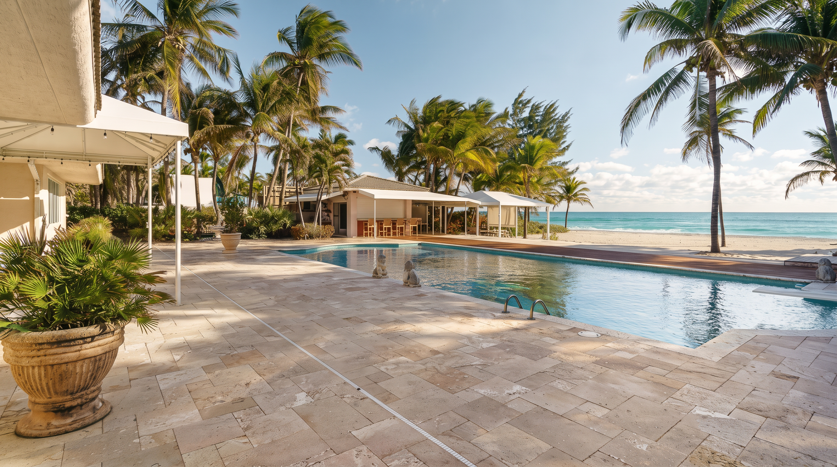 This expansive outdoor living space features a large, rectangular swimming pool set against a backdrop of lush palm trees and the ocean. A wide stone-paved patio surrounds the pool, leading to a covered outdoor bar area and a sandy beach just beyond the property line. The scene captures a serene, tropical atmosphere, emphasizing the seamless transition between luxury poolside relaxation and direct beach access.