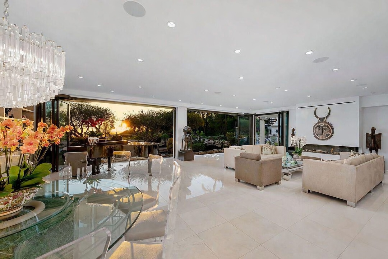 This expansive, open-concept living and dining area features polished light-colored flooring and a neutral color palette that enhances the sense of space. Large, retractable glass walls open the room to a sunset view of the garden, while a contemporary fireplace with a tall, minimalist surround serves as a focal point. The room is furnished with comfortable seating, a grand piano, and a glass-topped dining table, creating a sophisticated and airy atmosphere.