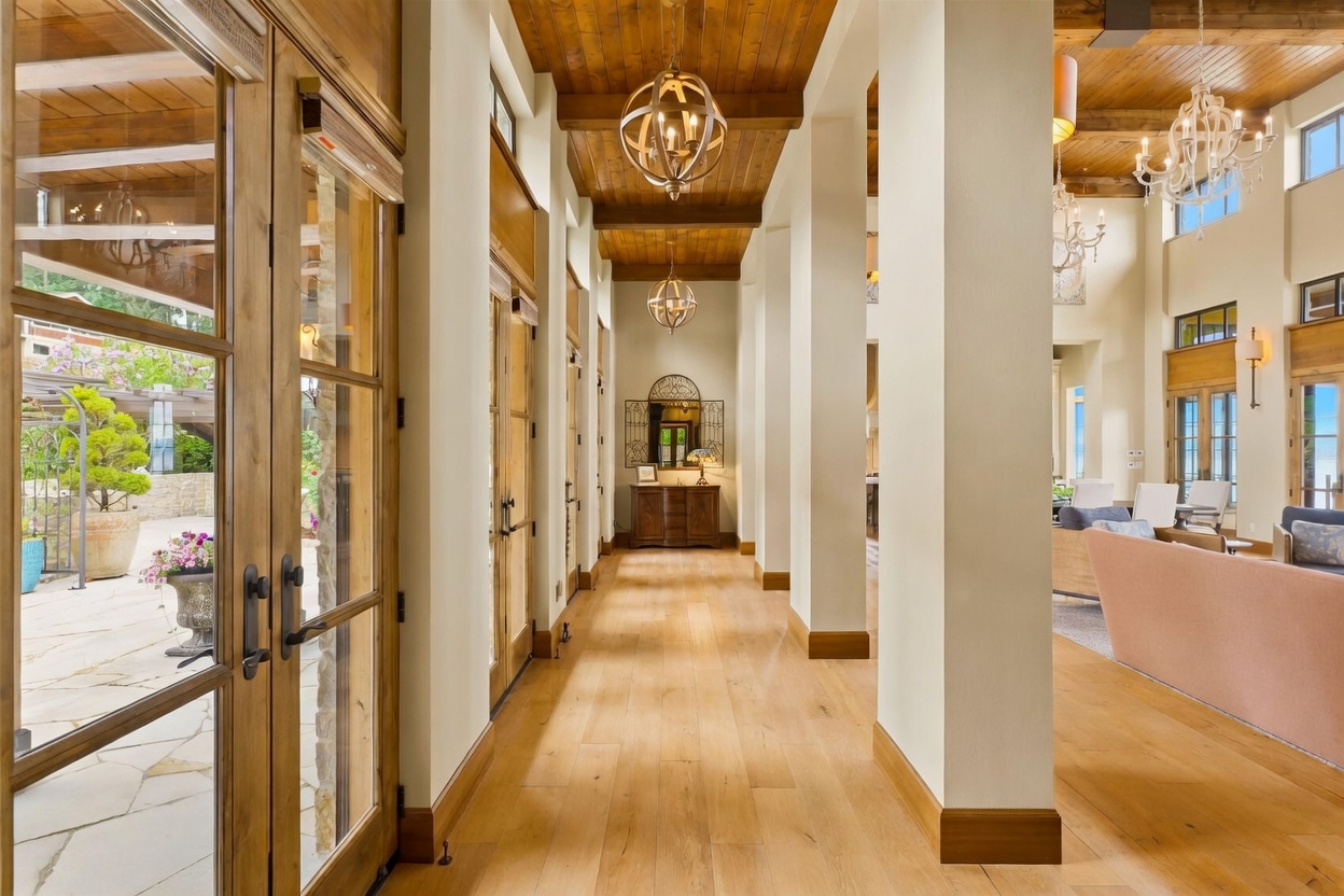 This elegant hallway features light wood flooring and a warm, rustic wood-paneled ceiling accented by two spherical pendant lights. The space is defined by tall, white structural columns that separate the corridor from an adjacent living area, creating an open and airy transition. A console table with a mirror and lamp sits at the end of the hall, while glass-paned doors on the left provide a view of the outdoor patio.