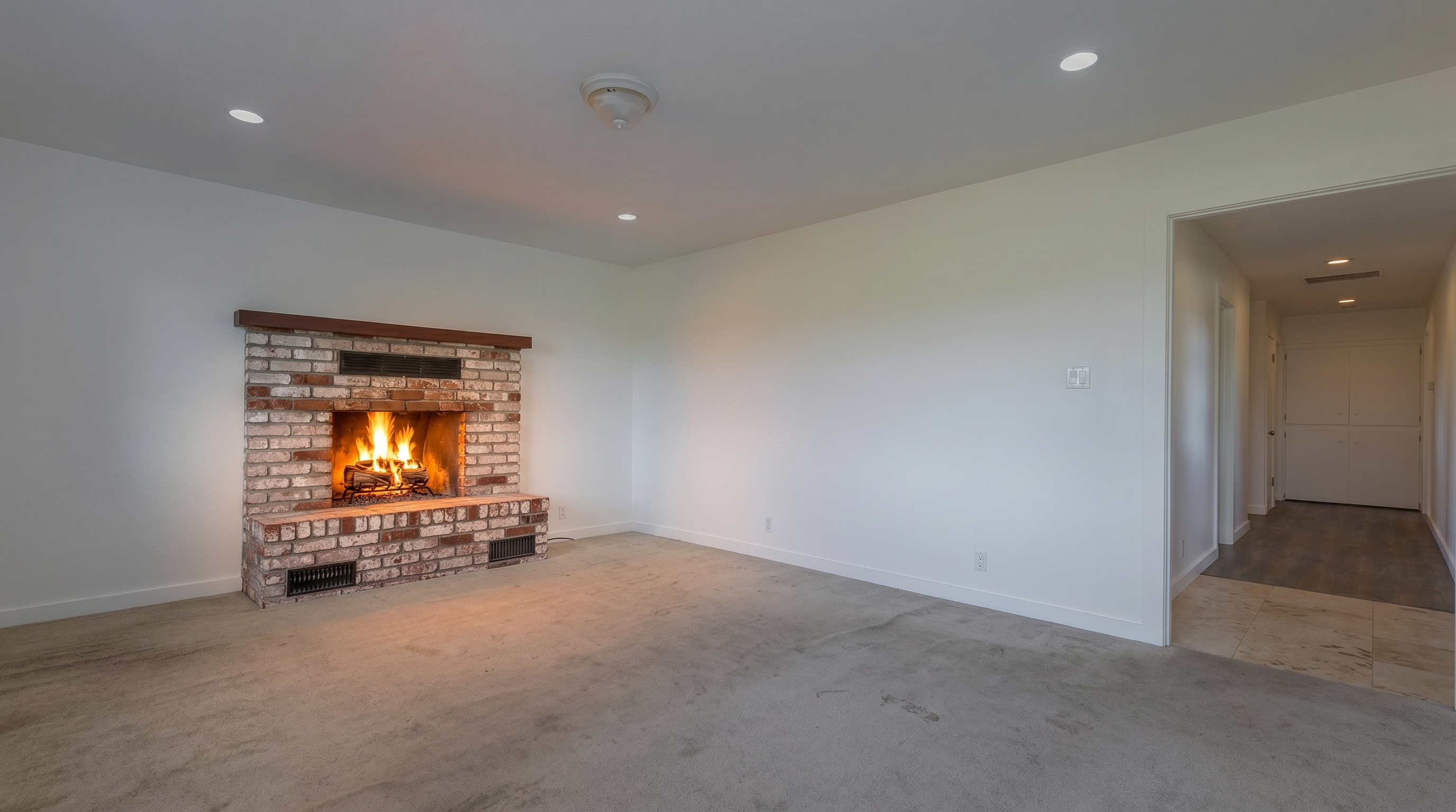 This spacious living room features neutral white walls and wall-to-wall carpeting, providing a blank canvas for personalization. A prominent brick fireplace with a wooden mantel serves as the focal point of the room, adding a touch of traditional character. The perspective looks toward the fireplace, with an open doorway on the right leading into a hallway with tiled flooring.