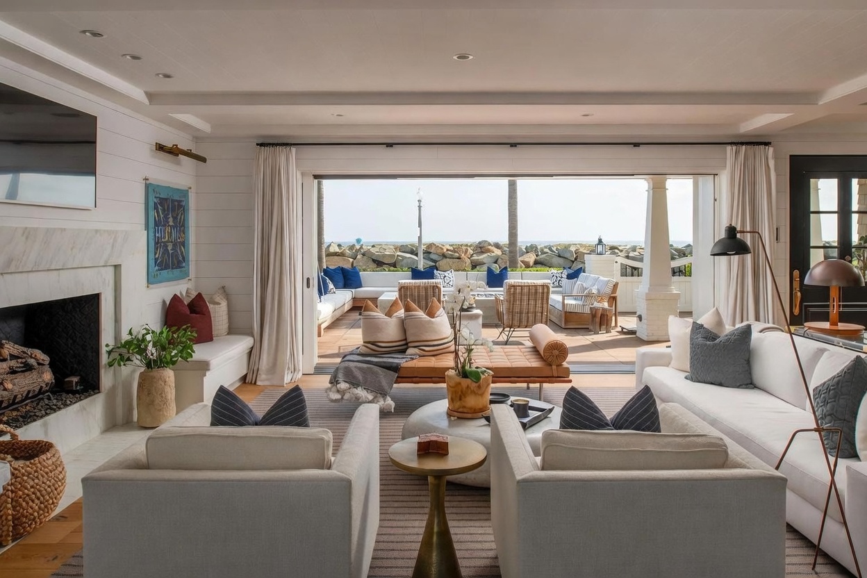 This bright and airy living room features a sophisticated coastal aesthetic with white shiplap walls and a large stone-clad fireplace surround. The space is anchored by a neutral-toned rug, plush seating, and a leather daybed, all oriented toward a wide opening that seamlessly connects the interior to an outdoor patio with ocean views. The open-concept design and natural light create an inviting, luxurious atmosphere perfect for relaxed seaside living.