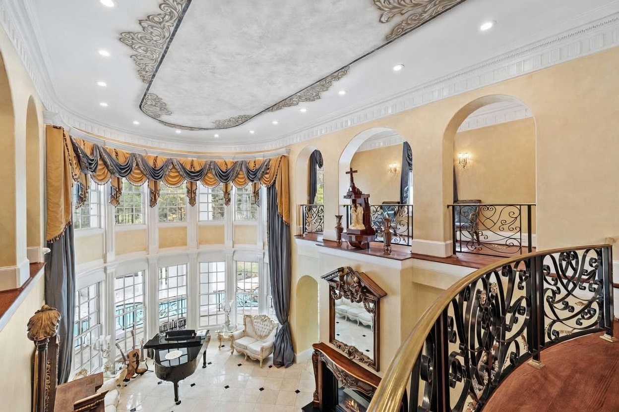 This grand, two-story living area features a dramatic curved wall of tall windows draped in elegant, multi-layered curtains. The space is defined by a sweeping staircase with ornate metal railings, a decorative ceiling medallion, and a mezzanine balcony overlooking the main floor. A grand piano sits on the polished light-toned tile floor, contributing to the opulent and formal atmosphere of the room.