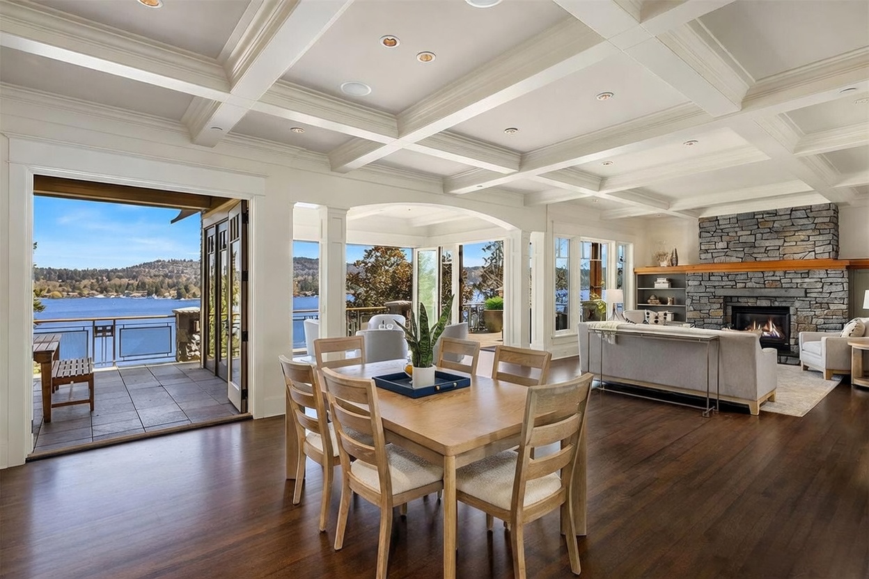 This expansive open-concept living and dining area features a coffered ceiling, rich hardwood flooring, and a striking floor-to-ceiling stone fireplace. Large glass doors open onto a spacious deck, seamlessly blending indoor and outdoor living while providing panoramic views of the lake. The space is elegantly furnished, creating a bright, airy, and sophisticated atmosphere perfect for entertaining.