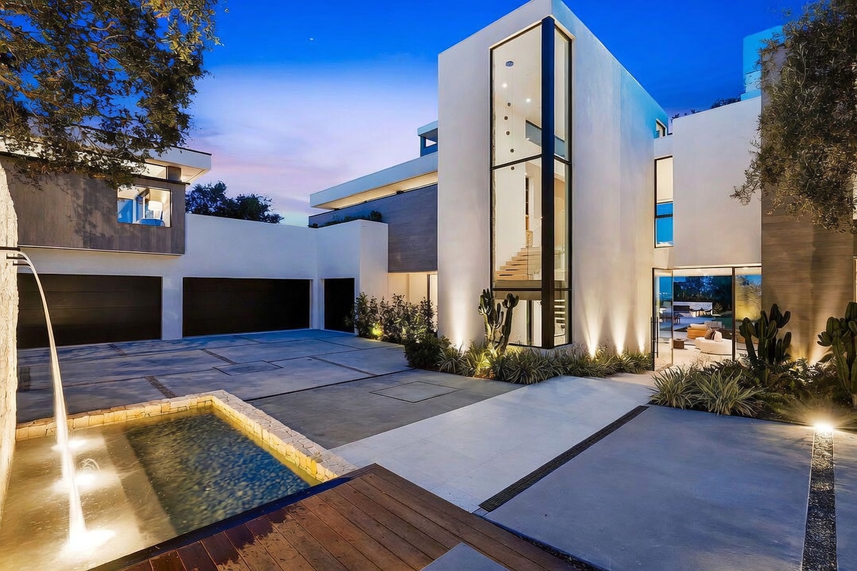 This striking modern residence features a dramatic, multi-story glass tower that serves as a focal point for the entryway. The exterior is characterized by clean, geometric lines, a neutral color palette, and a spacious paved motor court with an integrated water feature. Warm accent lighting highlights the architectural textures and landscaping, creating a sophisticated and inviting atmosphere at dusk.