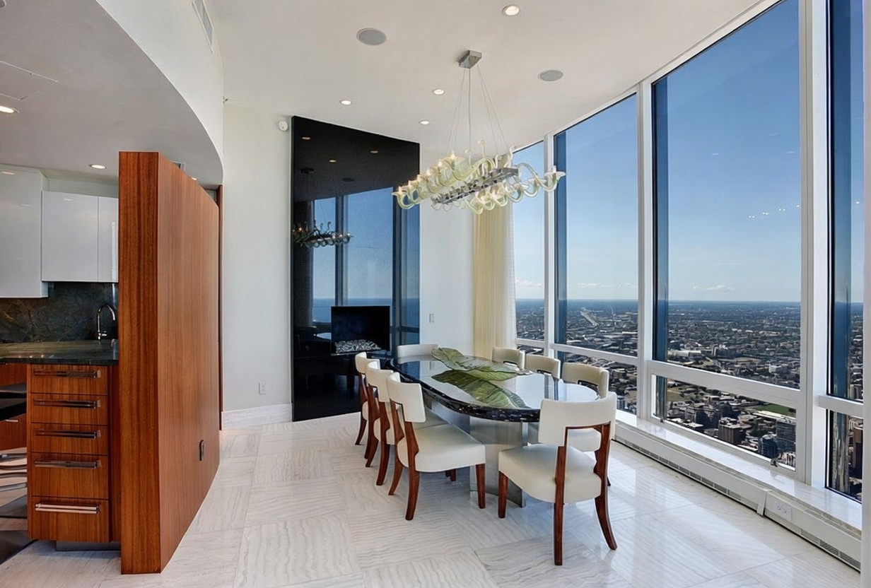 This modern dining room features a sleek oval table surrounded by six upholstered chairs, positioned beneath a decorative glass chandelier. The space is defined by expansive floor-to-ceiling windows that offer a panoramic view of the city skyline. A dark, reflective panel serves as a backdrop, while a wood-paneled wall separates the dining area from the adjacent kitchen, creating a sophisticated and open atmosphere.
