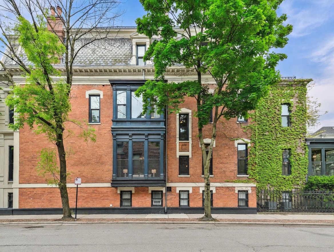 This classic brick townhouse features a prominent dark-painted bay window structure that adds architectural depth to the facade. The building is framed by mature trees and includes a section covered in lush green ivy, contributing to its established, historic charm. The perspective is a straight-on street view, capturing the symmetry of the windows and the detailed masonry work.