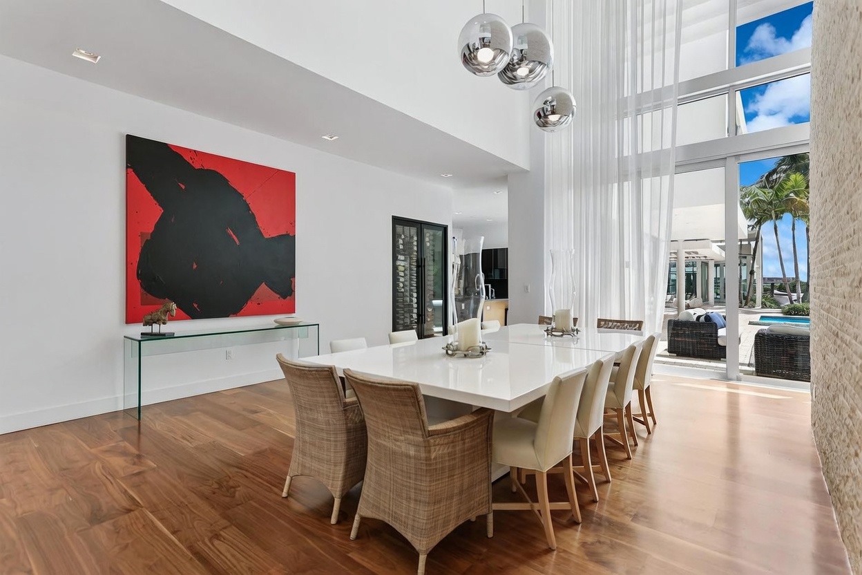 This bright and airy dining room features a large, glossy white table surrounded by woven-textured chairs, set against a backdrop of high ceilings and a striking abstract red and black painting. Large glass doors provide a seamless transition to the outdoor patio and pool area, allowing for abundant natural light. The space is accented by modern spherical pendant lights and warm wood flooring, creating a sophisticated and open atmosphere.