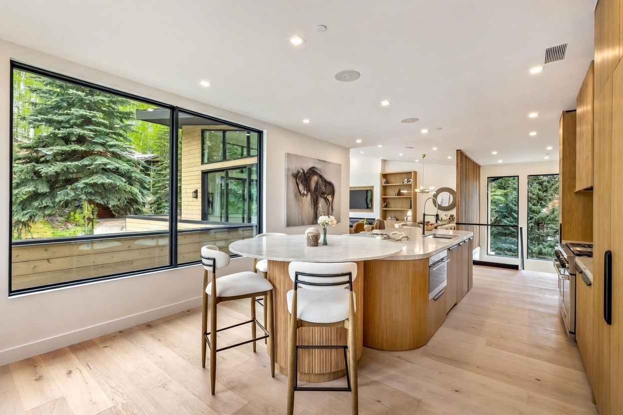 This modern, open-concept kitchen features a large, curved island with a light-toned stone countertop and integrated seating, complemented by light wood cabinetry. Large windows provide an expansive view of the surrounding forest, bathing the space in natural light. The design is sleek and minimalist, with a seamless transition into the adjacent living area, creating an airy and sophisticated atmosphere.
