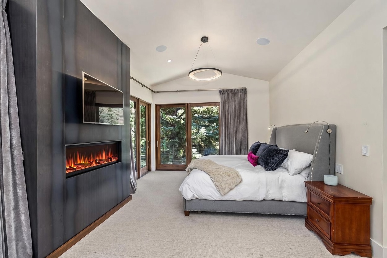 This primary bedroom features a contemporary design with a prominent dark metal-clad accent wall housing a built-in electric fireplace and a wall-mounted television. A plush gray upholstered bed serves as the focal point, complemented by a wooden nightstand and soft, neutral carpeting. Large glass doors provide access to an outdoor balcony, allowing natural light to fill the room while offering views of the surrounding trees.
