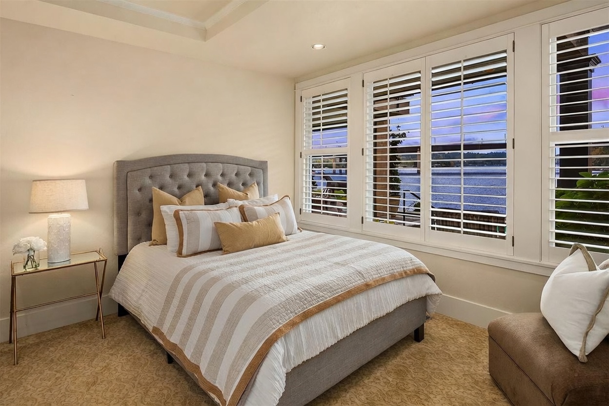 This cozy bedroom features a tufted gray upholstered headboard and a bed dressed in neutral-toned striped bedding. Large white plantation shutters cover the windows, which offer a scenic view of the water and a bridge outside. A small glass-topped side table with a lamp and a decorative floral arrangement sits to the left, creating a serene and inviting atmosphere.