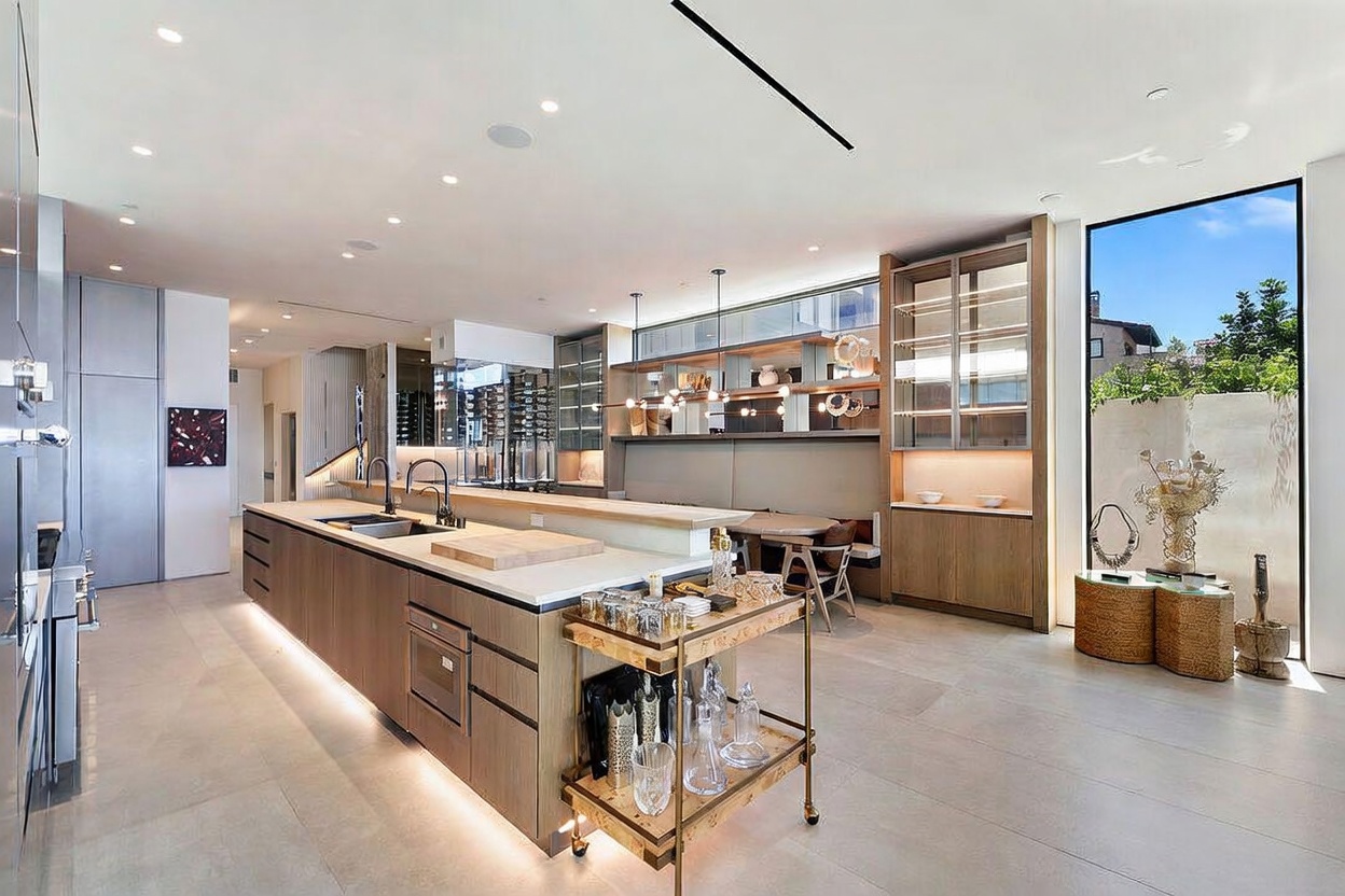 This expansive, modern kitchen features a large central island with a light-toned wood base and a stone countertop, complemented by a rolling bar cart in the foreground. The space includes integrated cabinetry, a built-in wine display, and a dining nook with a banquette, all illuminated by recessed lighting and a contemporary chandelier. A tall window provides natural light and a view of the outdoor area, creating a bright and sophisticated atmosphere.