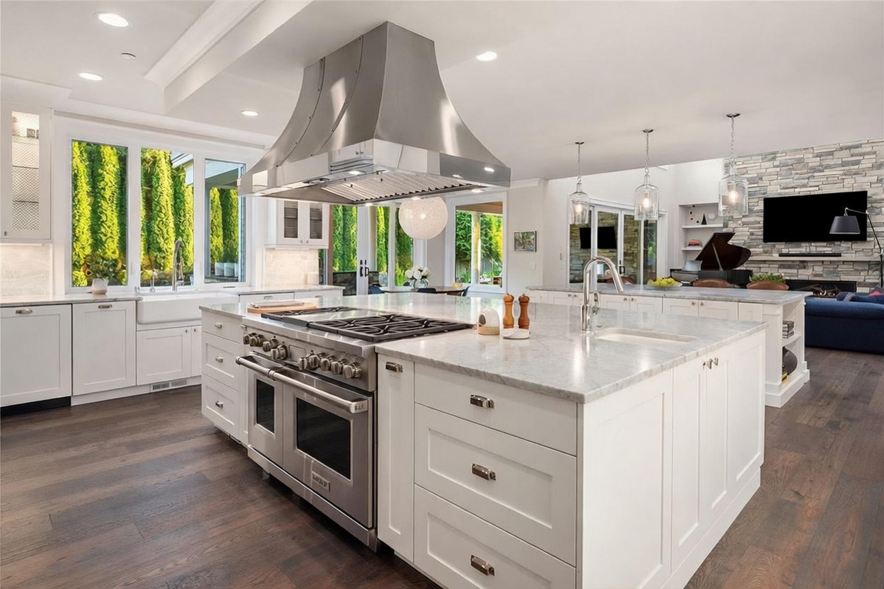 This expansive, high-end kitchen features a large central island with a stone countertop, a professional-grade stainless steel range, and a prominent stainless steel vent hood. The space is brightened by white cabinetry, dark wood flooring, and large windows that provide a view of the lush greenery outside. In the background, a living area with a stone-clad chimney breast and a grand piano creates an open-concept flow, perfect for entertaining.