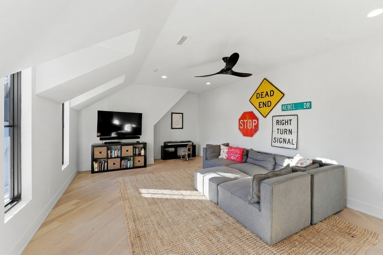 This spacious attic-style living room features light wood flooring, white walls, and a vaulted ceiling with a modern black ceiling fan. A large gray sectional sofa sits atop a textured area rug, facing a media console with a television and built-in storage. The wall is decorated with vintage-style road signs, adding a playful, casual aesthetic to the bright, open space.