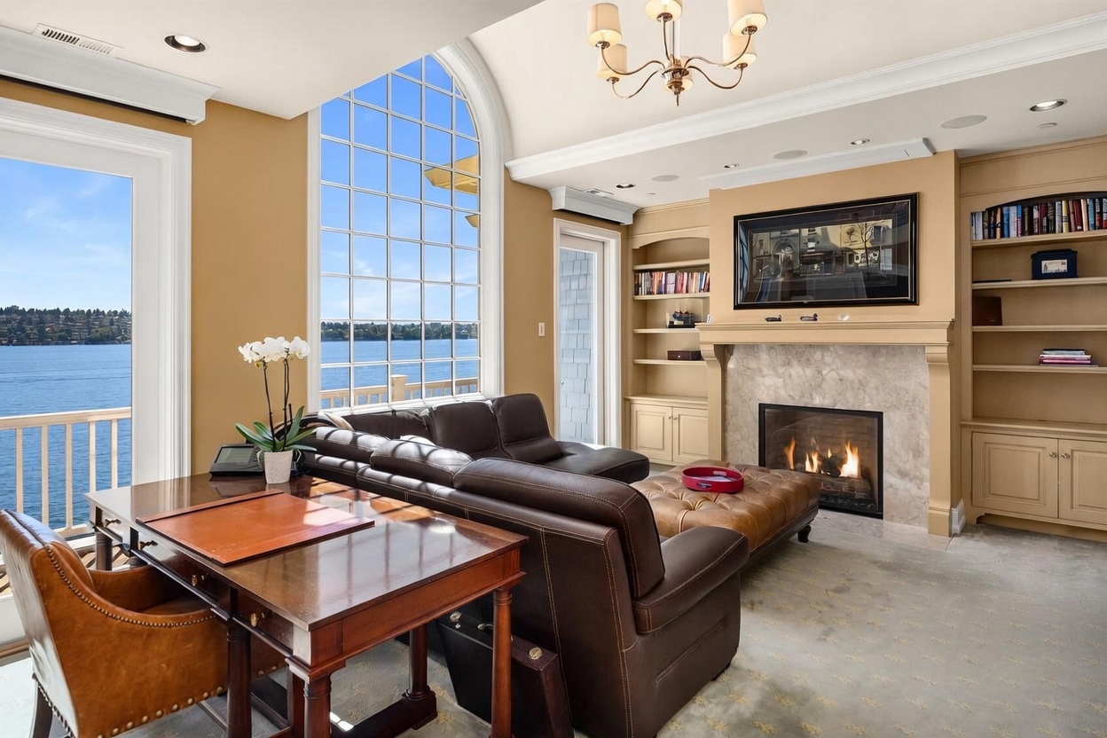 This elegant home office or study features a rich, dark wood desk, a plush leather sofa, and a matching ottoman, all arranged to face a cozy fireplace with a built-in bookshelf surround. A massive, arched floor-to-ceiling window provides a breathtaking, panoramic view of the lake, flooding the room with natural light. The space exudes a sophisticated, tranquil atmosphere, perfect for both focused work and relaxation.