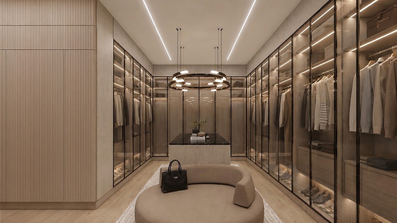 This luxurious walk-in closet features floor-to-ceiling glass-front cabinetry with integrated lighting, providing a sophisticated display for clothing and accessories. A central island with a dark stone top serves as a focal point, complemented by a curved, upholstered bench in the foreground. The space is illuminated by a modern circular chandelier and recessed linear lighting, creating an elegant and high-end atmosphere.