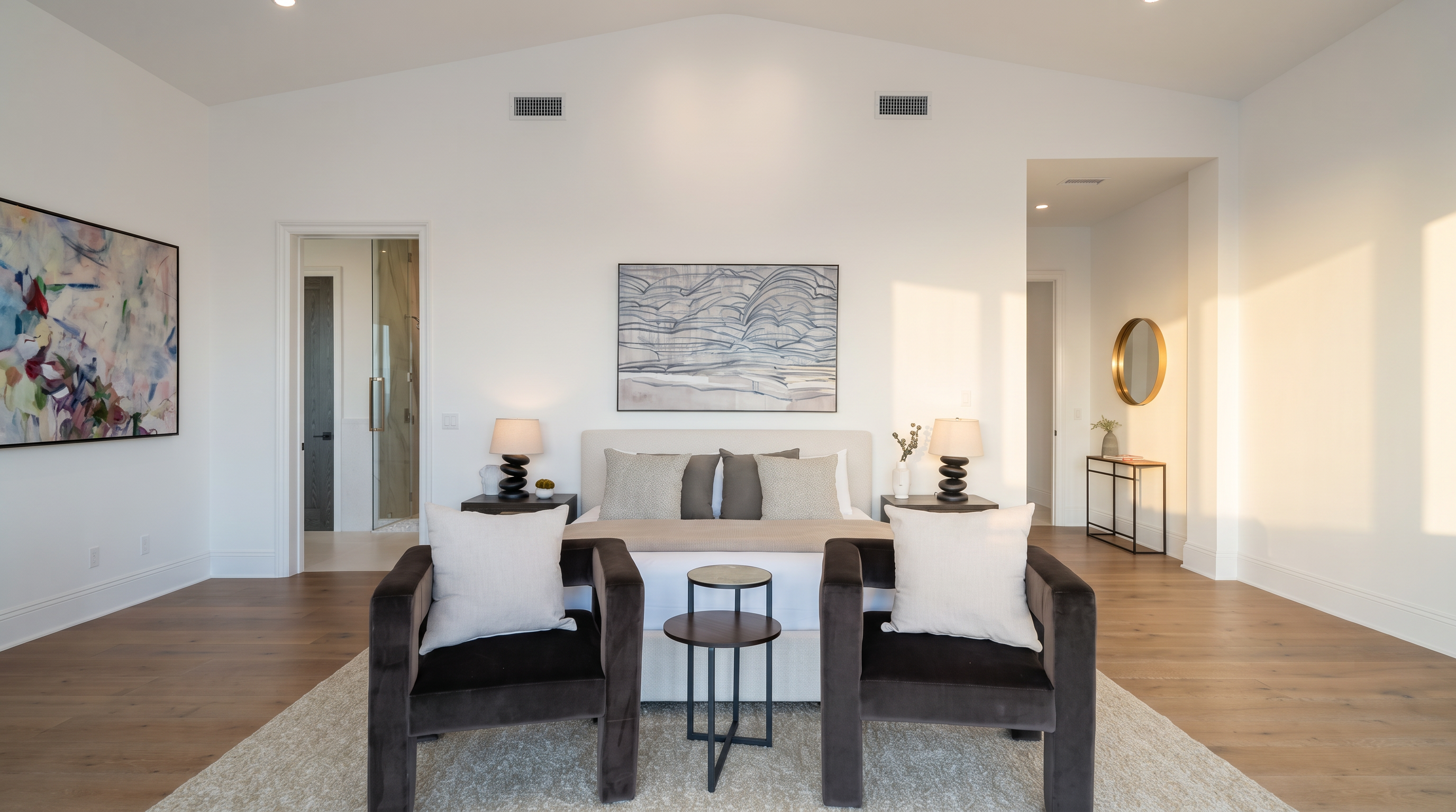 This spacious primary bedroom features a vaulted ceiling and light wood flooring, creating an airy and sophisticated atmosphere. A central bed is flanked by two matching nightstands with lamps, while two dark-upholstered armchairs sit in the foreground with a small round side table between them. The room is decorated with neutral tones and a large piece of abstract wall art, providing a clean and modern aesthetic.