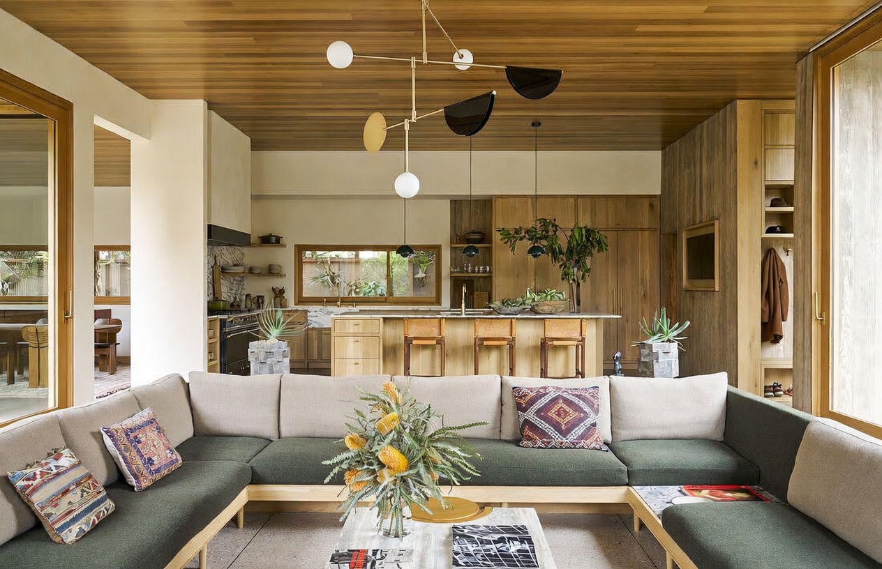 This open-concept living area features a large, U-shaped sectional sofa with dark green cushions and light wood framing, centered around a low coffee table. The space flows seamlessly into a kitchen area with a central island, light wood cabinetry, and a distinctive modern chandelier hanging from a warm-toned wood-paneled ceiling. The design aesthetic is organic and contemporary, utilizing natural textures and a neutral color palette to create a cohesive, inviting atmosphere.
