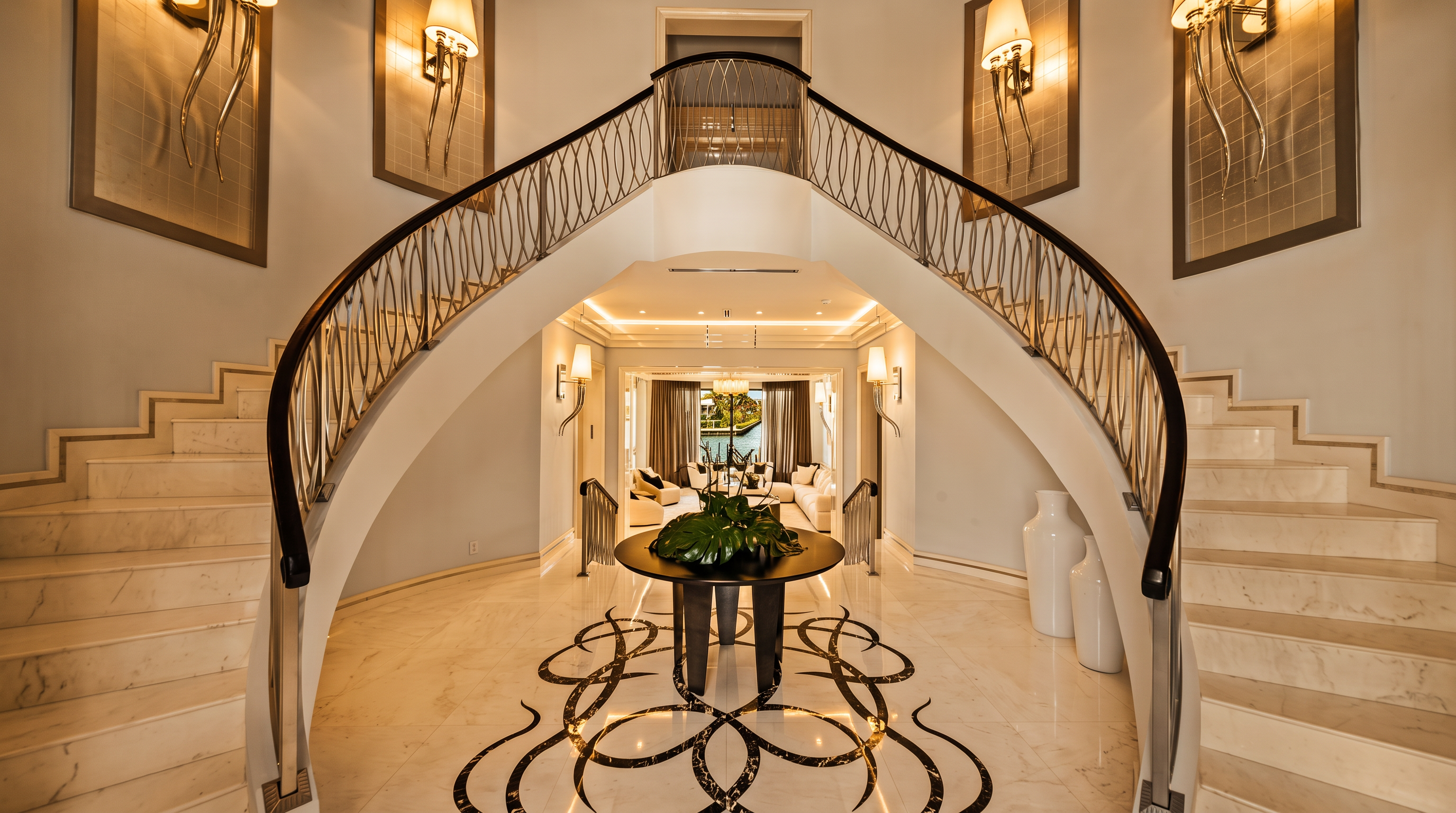 This grand foyer features a dramatic, symmetrical double staircase with intricate metal railings that curve upward from a central landing. The polished stone floor is adorned with a large, ornate dark inlay pattern, centered beneath a round pedestal table holding a lush green plant. The perspective looks directly through the archway toward a bright living area with views of the water, creating a sense of openness and luxury.