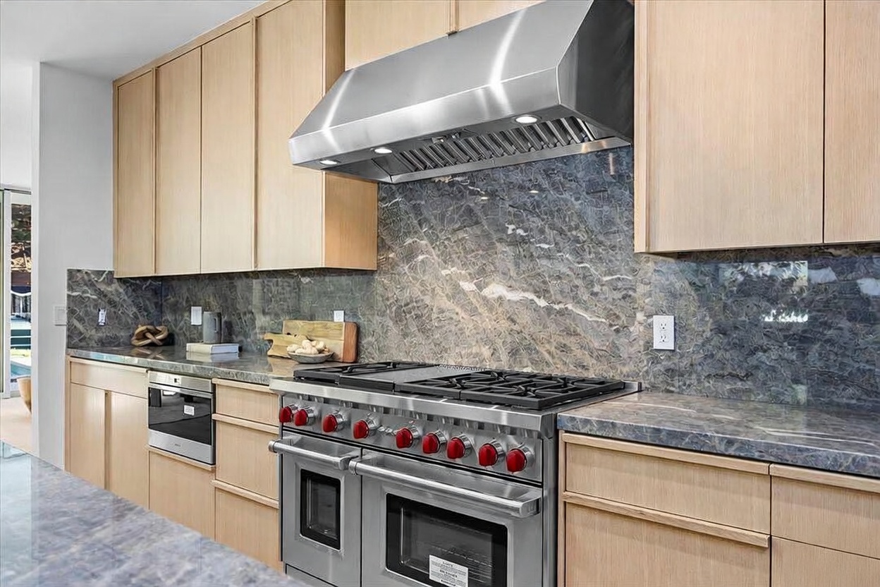 This modern kitchen features light wood cabinetry paired with a striking dark, veined stone backsplash and matching countertops. A professional-grade stainless steel range with a matching vent hood serves as the focal point, complemented by an integrated oven to the left. The clean lines and neutral color palette create a sophisticated and functional culinary space.